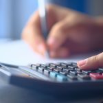 Mãos usando uma calculadora e fazendo anotações para organizar o orçamento doméstico.