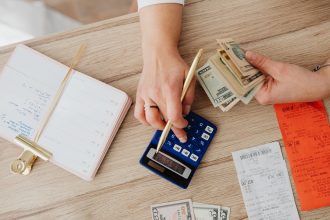 Mãos organizam recibos, dinheiro e uma calculadora sobre uma mesa de madeira.