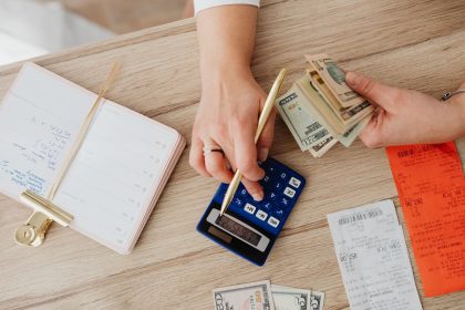 Mãos organizam recibos, dinheiro e uma calculadora sobre uma mesa de madeira.